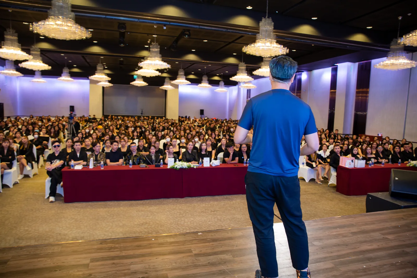 Hội trường tiệc kín bàn kín người tham dự, người hướng dẫn đứng chia sẻ phía trước dưới đèn chùm trần